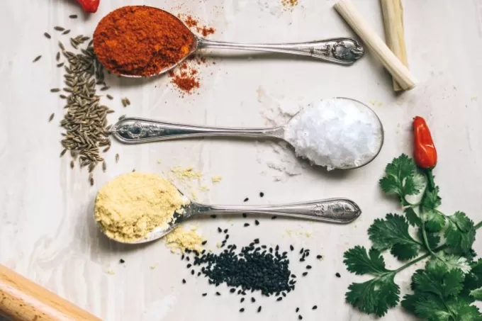 Three silver tablespoons holding an asssortment of spices on a table next to parsle leaves, red chili pepper, and lemongrass