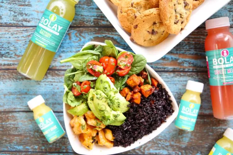 Bolay spicy thai shrimp bowl with forbidden black rice, a platter of cookies, and an assortment of freshly squeezed juices on a table