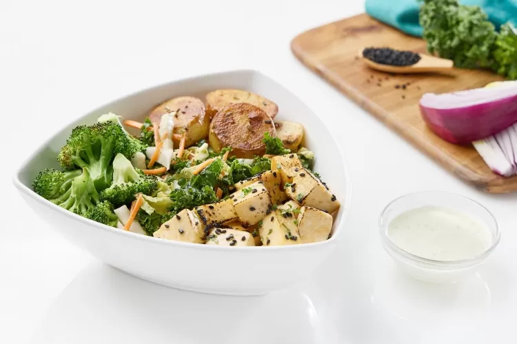 Bolay miso glazed tofu bowl with marinated tofu, roasted potatoes, kale salad, and garlic broccoli next to a cup of garlic sauce and a cutting board with kale, whole peppercorns, and a quarter of a re