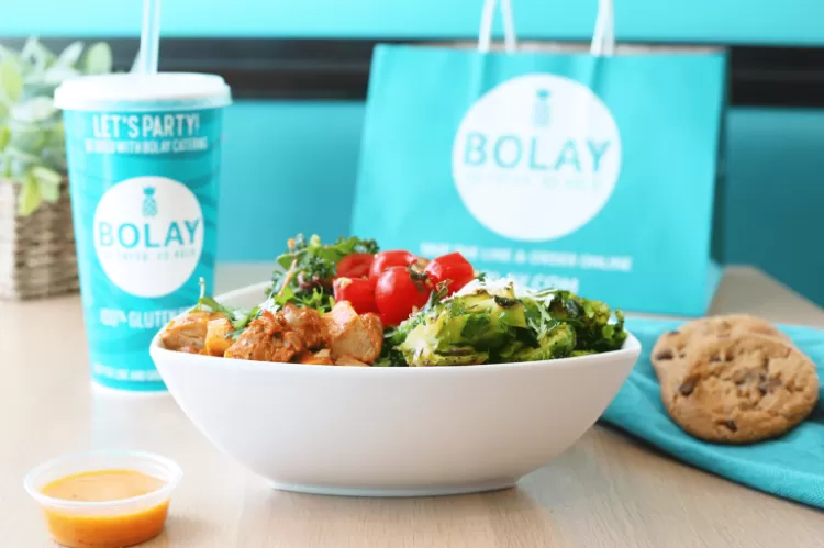 A table inside a Bolay restaurant with a teriyaki chicken bowl, a cup of habanero sauce, a fountain drink, two cookies, and a blue Bolay takeout bag