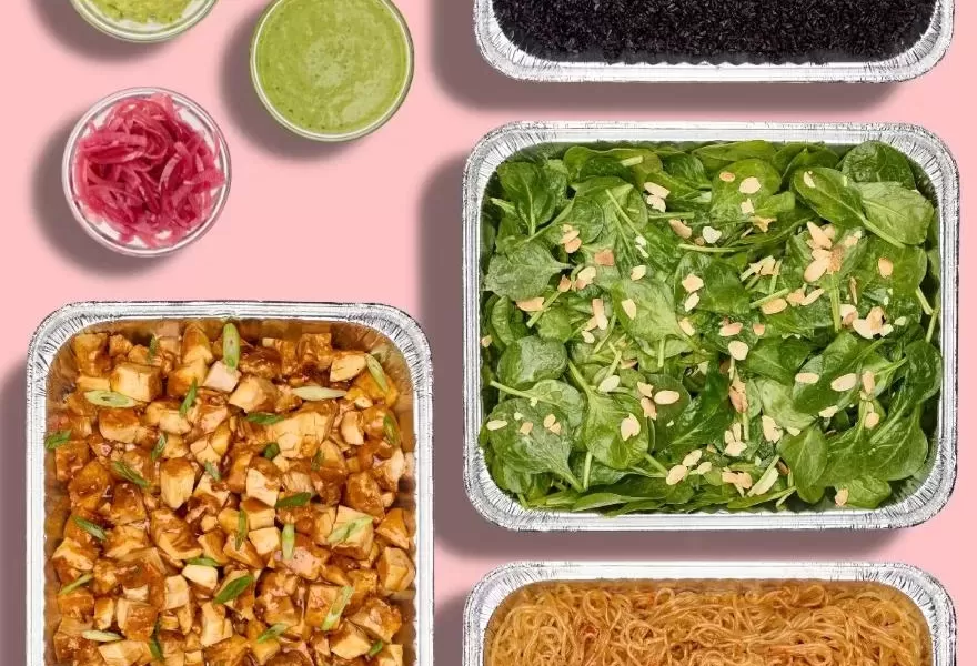 Bolay Family Meal trays containing teriyaki chicken, forbidden black rice, asian sweet potato noodles, and spinach and almond salad with bowls of fresh avocado, pickled red onion, and pesto on the sid